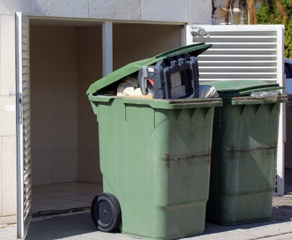 A large green wheelie bin with a black lid, positioned on a paved driveway outside a residential building. The bin is filled with assorted rubbish, including plastic bags and various waste materials, some of which are spilling slightly over the top. The textured surface of the bin shows weathering and minor dirt marks. Behind the bin, there is a small outdoor structure or enclosure with tiled flooring and a partial wall, possibly part of a garage or storage area. To the right, a second waste bin with a similar design is partially visible. The scene is set during daylight, with natural light highlighting the outdoor environment. This image illustrates typical rubbish collection and disposal in the Ruislip area, aligning with services provided by Business Waste Removal Ruislip for efficient waste management and clearance of household or commercial rubbish.