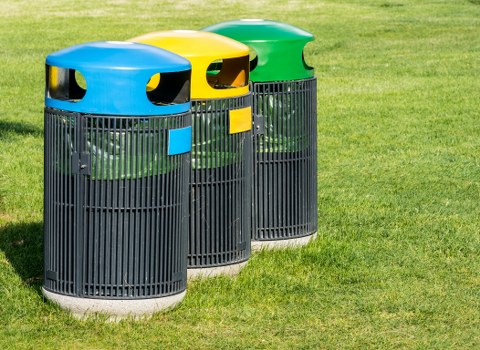 Three cylindrical outdoor rubbish bins positioned on a well-maintained grassy area, each with a black plastic slatted body and a smooth, curved plastic lid in bright blue, yellow, and green respectively. The lids have small openings for waste disposal and are fitted with matching colour-coded labels on the front. The bins are aligned side by side, with the blue one closest to the camera, followed by yellow and green, all standing on a paved or concrete base. The background shows lush, green grass with no other objects or structures visible, suggesting a park, communal outdoor space, or a residential area. The lighting indicates natural daylight, highlighting the vivid colours of the lids against the darker tone of the bodies, consistent with an environment where waste removal services such as those offered by Business Waste Removal Ruislip are utilized for maintaining cleanliness and waste collection in local community settings.