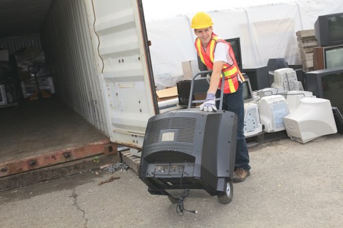 A worker wearing a yellow safety helmet, high-visibility vest, and work gloves is outdoors at what appears to be a storage or loading area, standing on a paved surface next to a partially open shipping container. The worker is holding a portable television set with a black plastic casing, a curved screen, and visible control buttons on the front, with the power cable dangling. Behind the worker, there are various other electronic appliances, such as old computer monitors and CRT televisions, stacked on the ground near the container entrance. The scene is set in natural daylight, with a white tent or temporary structure visible in the background, and the environment resembles a waste collection or disposal site for electronic waste, aligning with rubbish removal services offered by Business Waste Removal Ruislip in the area.
