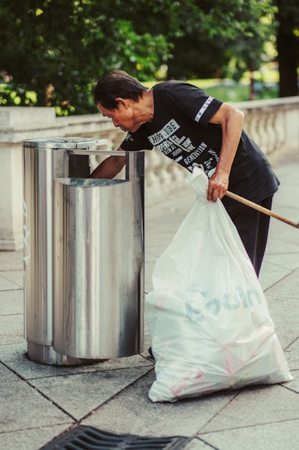 A man with dark hair and a dark T-shirt is seen bending over to dispose of rubbish into a metallic, cylindrical outdoor waste bin with a slot on top, located on a paved area. The T-shirt has a graphic