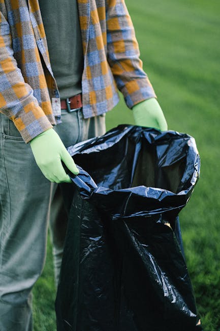 A man with dark hair and a dark T-shirt is seen bending over to dispose of rubbish into a metallic, cylindrical outdoor waste bin with a slot on top, located on a paved area. The T-shirt has a graphic