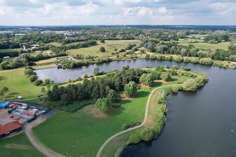 An aerial view of Ruislip Lido featuring a large, irregularly shaped body of water encircled by a narrow, winding pathway lined with lush, green trees and shrubs. The water appears calm and reflective