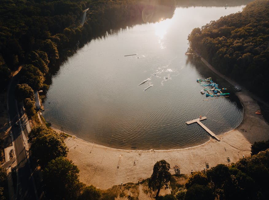 An aerial view of Ruislip Lido featuring a large, irregularly shaped body of water encircled by a narrow, winding pathway lined with lush, green trees and shrubs. The water appears calm and reflective